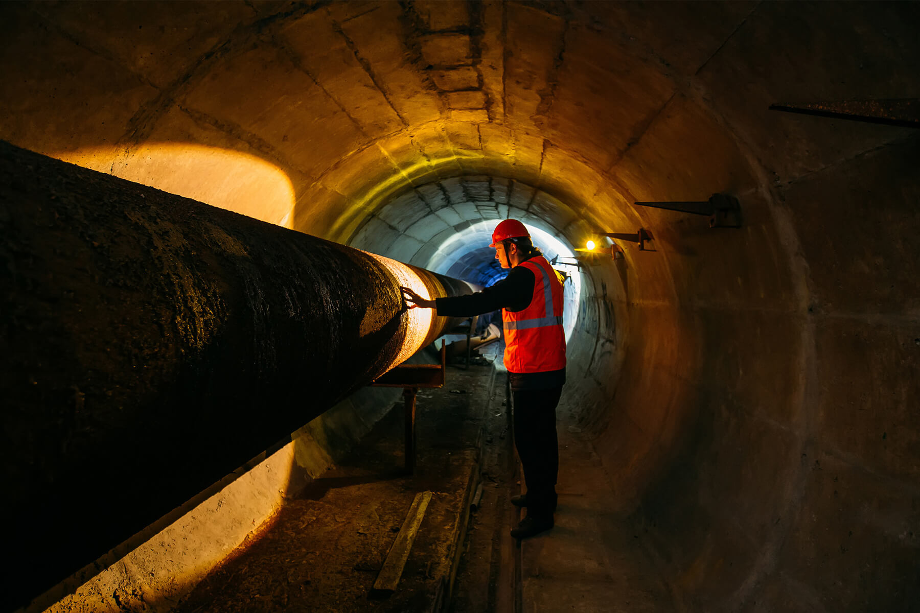 Eine Person mit Schutzweste und Helm steht in einem dunklen Tunnel. Er beleuchtet mit einer Arbeitslampe einen Teil eines Rohres und betrachtet diesen.