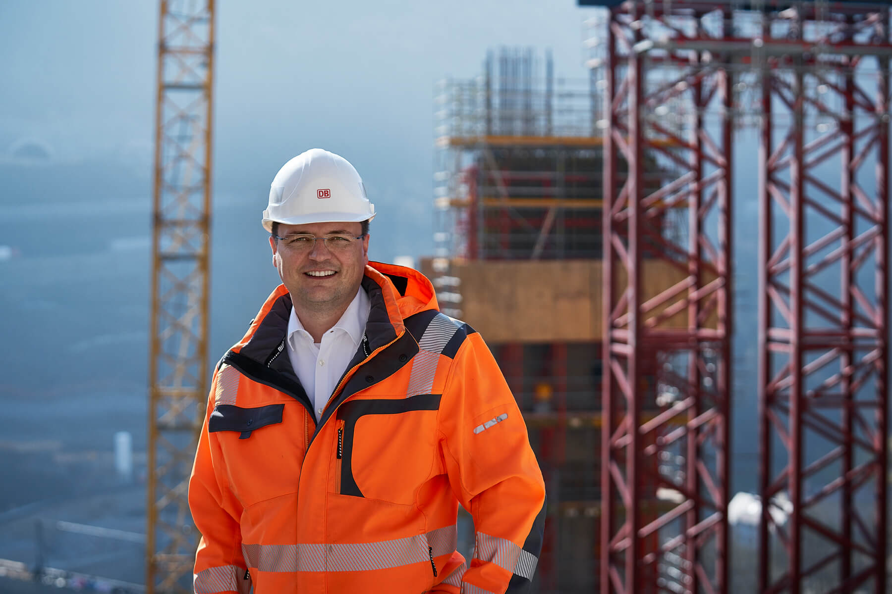 Ein Mann mit Neonfarbener Jacke und Schutzhelm lächelt in die Kamera. Im Hintergrund ist eine Baustelle zu sehen mit Kränen.