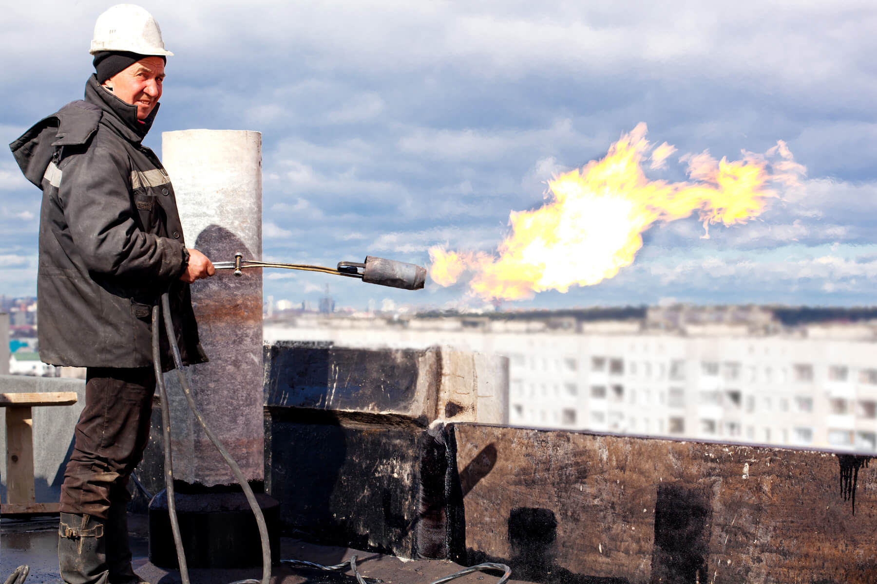 Eine Person mit Helm und Schutzkleidung hält einen Gasbrenner der gerade eine Flamme erzeugt und steht auf einem Hausdach.