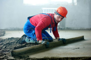 Ein Mann streicht Beton glatt. Er hat eine blaue Latzhose und einen Roten Helm an. Für die Arbeit kniet er auf dem Boden.
