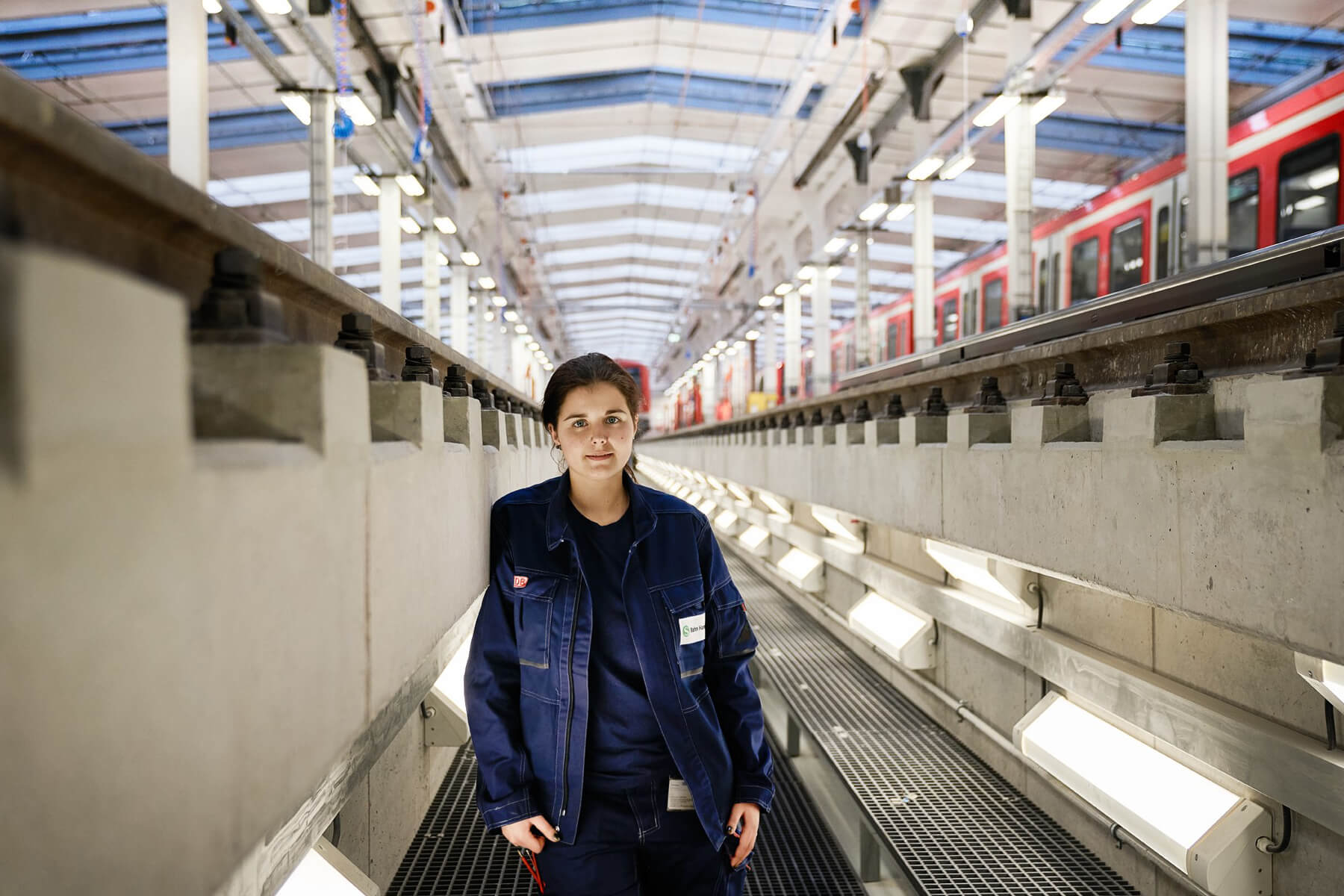 Eine Frau steht in einer Halle mit Zügen im Bereich unterhalb der Gleis. Die Halle ist mit vielen großen Lampen im Decken- und Bodenbereich ausgestattet.