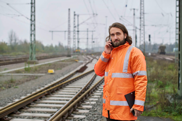 Ein Mann in einer orangenen Warnjacke steht an der Gleisstrecke. Er telefoniert mit dem Handy und hält in der anderen Hand ein Tablet.