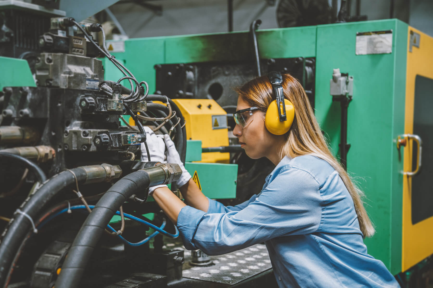 Eine Frau mit Handschuhen und Gehörschutz arbeitet an einer großen Maschine mit vielen Kabeln.
