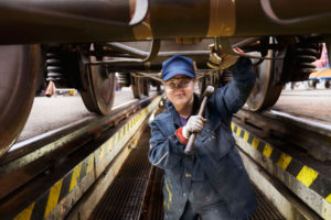 Eine Frau steht mit stark verschmutzter Arbeitskleidung unter einem Zug. In der Hand hält sie einen Metallhammer.