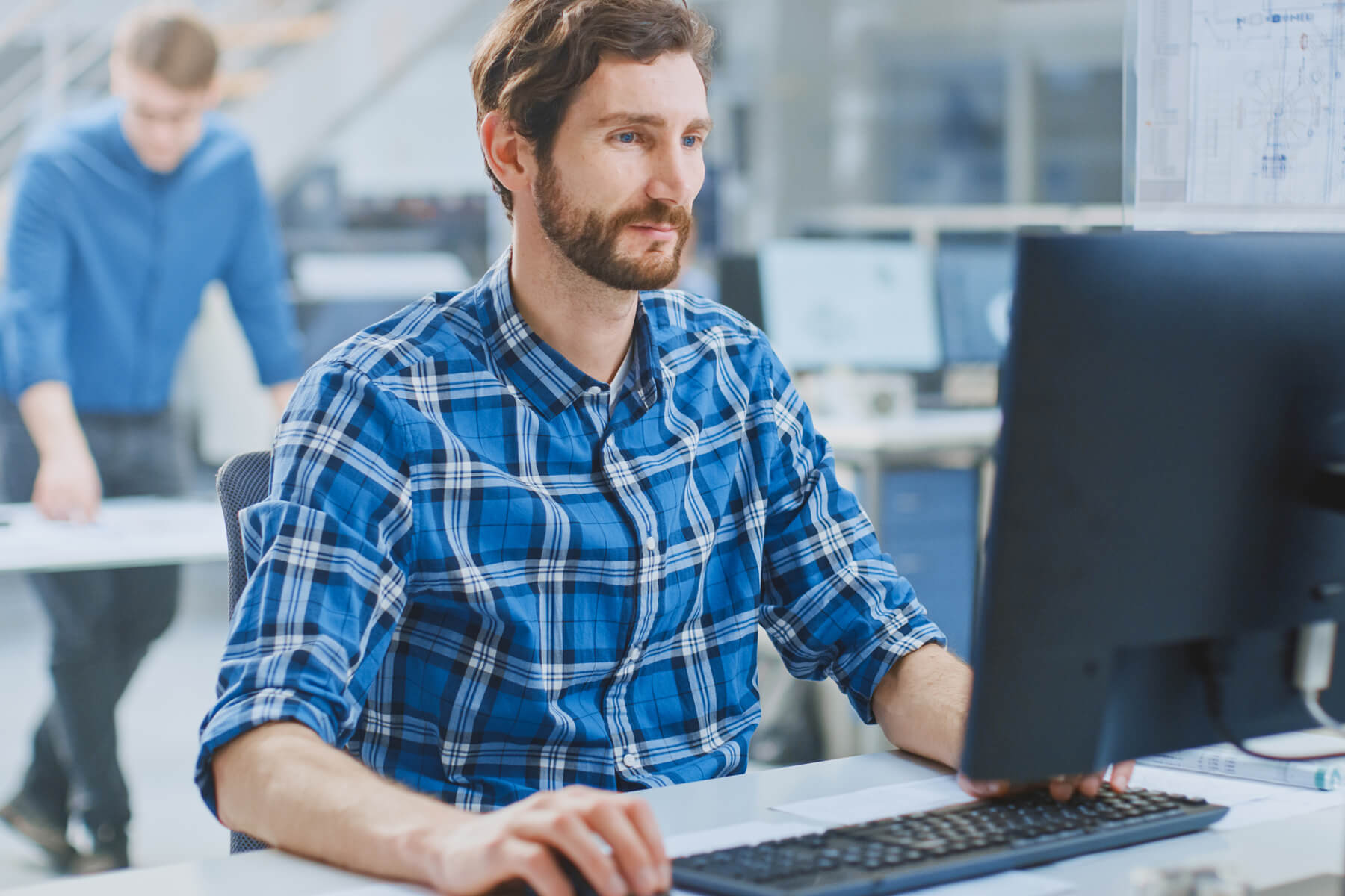Ein Mann sitz an einem Schreibtisch vor einem Bildschirm und tippt auf der Tastatur. Im Hintergrund ist ein Büro zusehen.