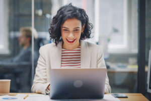 Eine Frau sitzt vor ihrem Laptop und lächelt. Im Hintergrund ist ein Büro zu sehen.